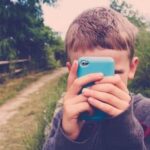 photograph of a young boy in a dark top with light brown hair holding a phone close t his face as though taking a photo